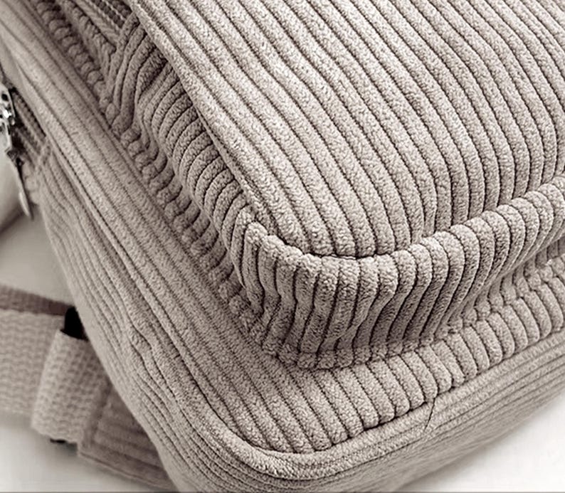 Puede incluir: Primer plano de una mochila de pana beige. La tela tiene una textura acanalada, con l&iacute;neas verticales visibles. El dise&ntilde;o de la bolsa incluye una solapa y una esquina redondeada, lo que sugiere un estilo informal. El color es un marr&oacute;n claro neutro.