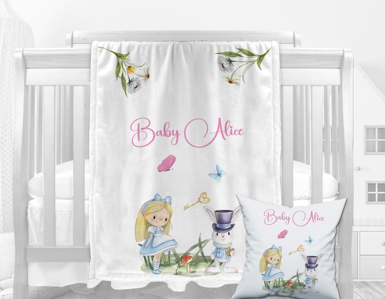 K&ouml;nnte beinhalten: Eine wei&szlig;e Babydecke mit einer Aquarell-Illustration von Alice im Wunderland-Charakteren, darunter Alice, der wei&szlig;e Hase und ein Schl&uuml;ssel. Die Decke tr&auml;gt den Text "Baby Alice" in rosa kursiver Schrift.