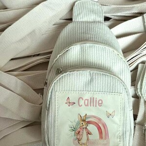 Puede incluir: Una mochila bandolera de pana gris claro con un compartimento principal con cremallera y un bolsillo delantero m&aacute;s peque&ntilde;o. El bolsillo delantero presenta un parche con el nombre "Callie", mariposas, un arco&iacute;ris y un conejo con abrigo. La bolsa tiene correas ajustables.
