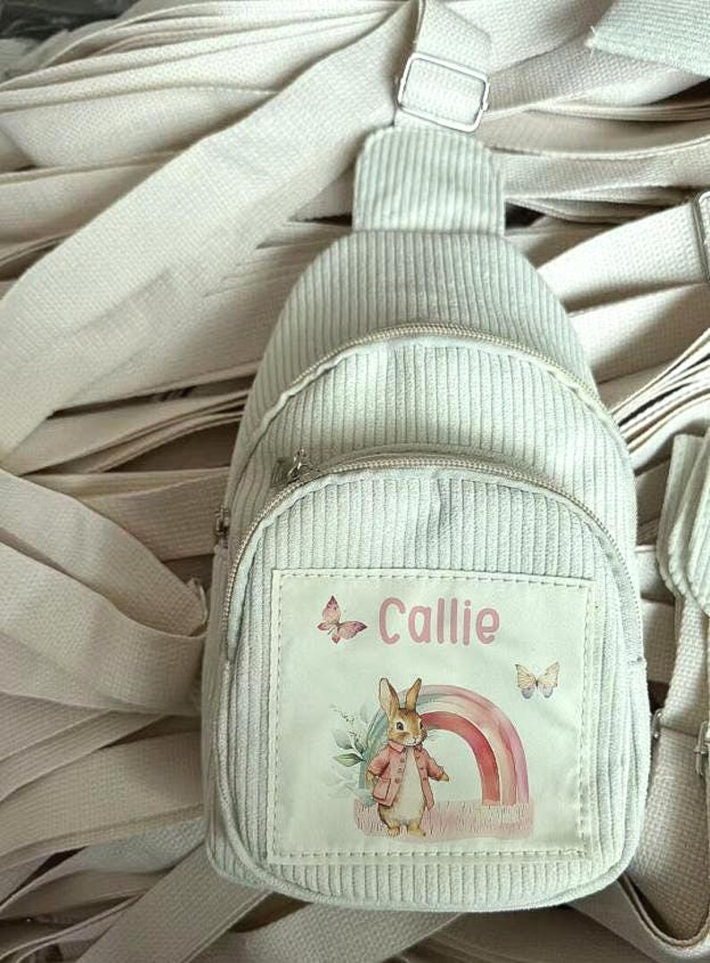 Puede incluir: Una mochila bandolera de pana gris claro con un compartimento principal con cremallera y un bolsillo delantero m&aacute;s peque&ntilde;o. El bolsillo delantero presenta un parche con el nombre "Callie", mariposas, un arco&iacute;ris y un conejo con abrigo. La bolsa tiene correas ajustables.