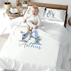 Puede incluir: Juego de cama blanco con un diseño de Peter Rabbit y el nombre "Arthur" en escritura azul. El juego incluye una funda nórdica y fundas de almohada. Un niño está sentado en la cama.