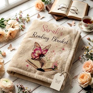 以下が含まれることがあります： 「Sarah's Reading Blanket」の文字と蝶のデザインが施されたベージュ色の読書用ブランケット。 ピンクと白の蝶と花のアクセントが施された開いた本が特徴です。 蝶の画像が付いたタグが取り付けられています。