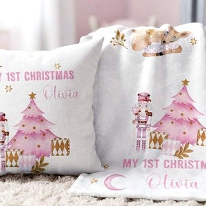 Puede incluir: Un cojín cuadrado blanco y una manta de forro polar blanca a juego con un árbol de Navidad rosa, un cascanueces, regalos y un cervatillo. Ambos artículos tienen impreso el texto "MY 1ST CHRISTMAS" y el nombre "Olivia" en letra cursiva rosa.