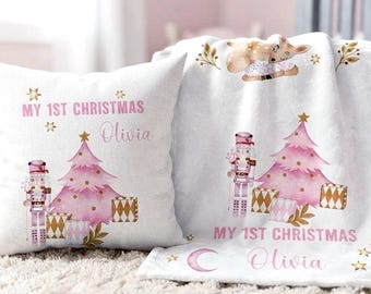 Könnte beinhalten: Ein weißes quadratisches Dekokissen und eine passende weiße Fleecedecke mit einem rosa Weihnachtsbaum, einem Nussknacker, Geschenken und einem Rehkitz. Beide Artikel sind mit dem Text "MY 1ST CHRISTMAS" und dem Namen "Olivia" in rosa Schrift bedruckt.