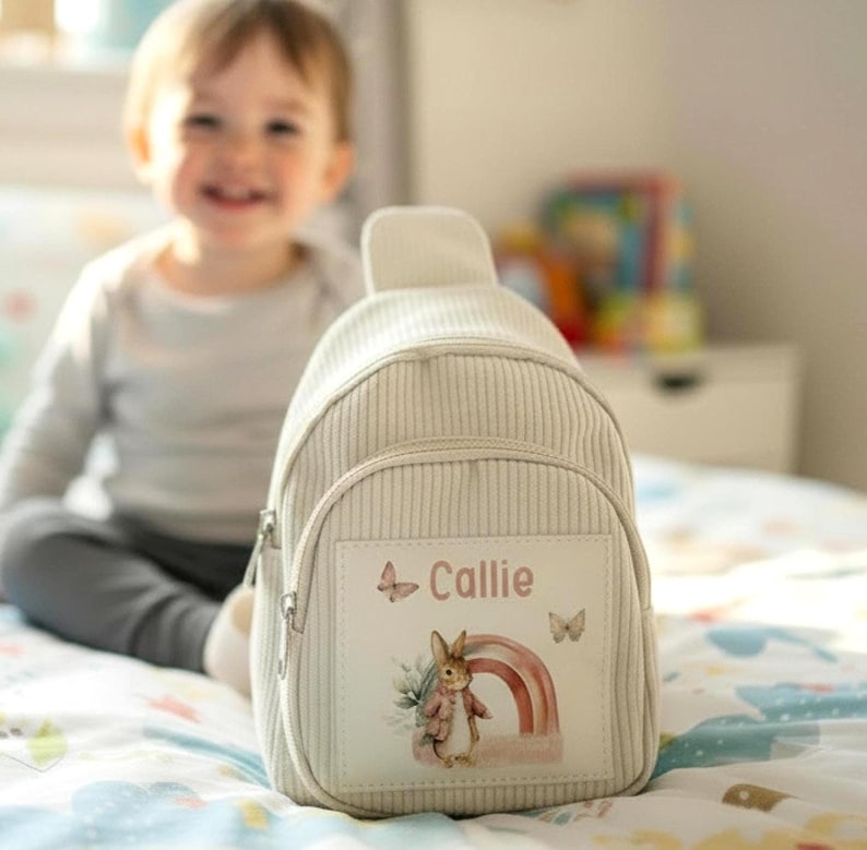 Puede incluir: Una mochila beige de pana con un bolsillo frontal con cremallera. La mochila presenta un parche rectangular con el nombre "Callie", una ilustraci&oacute;n de acuarela de un conejo, un arco&iacute;ris y mariposas. La mochila est&aacute; dise&ntilde;ada para un ni&ntilde;o.