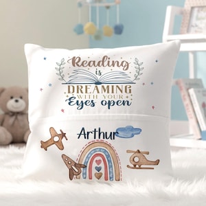 Könnte beinhalten: Ein weißes quadratisches Kissen mit dem Text Reading is DREAMING WITH YOUR EYES OPEN über einer Tasche. Die Tasche zeigt den Namen Arthur, eine blaue Wolke, ein Holzflugzeug, eine Holzrakete, einen Holzhelikopter und einen Regenbogen.