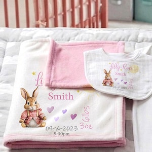 May include: A personalized baby gift set featuring a white and pink blanket, a pink blanket, and a bib. The blanket has a bunny illustration, name, birth date, time, and weight. The bib has a bunny illustration and name.