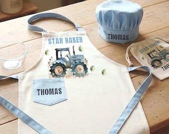 May include: A cream-colored apron with light blue straps and a pocket featuring the name THOMAS, a blue tractor graphic, and the text STAR BAKER. Includes a matching light blue chef hat with THOMAS printed on it and a small drawstring bag.