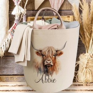 Peut inclure: Un panier à linge beige avec une illustration aquarelle d'une vache des Highlands. Le panier, avec une poignée, contient du linge plié. Le nom "Abbie" est écrit en dessous. L'arrière-plan comprend des planches de bois et des éléments décoratifs.