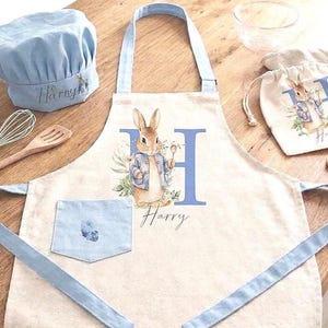 Pode incluir: Um avental, um chapéu de chef e um saco com cordão com um design de Peter Rabbit. O avental tem um bolso e alças azuis, com o nome "Harry" impresso no avental e no chapéu. A ilustração de Peter Rabbit segura uma colher.