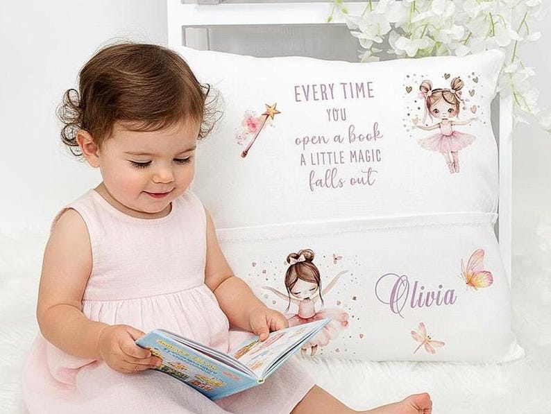 Op de afbeelding: Een wit kussen met een zak, versierd met een ballerina-ontwerp en de tekst "Every time you open a book a little magic falls out." Het kussen heeft ook de naam "Olivia" erop gedrukt. Een jong kind houdt een boek vast.