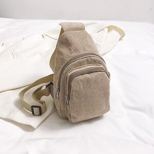 Puede incluir: Bolso bandolera de pana beige con un bolsillo frontal con cremallera y una correa ajustable. El bolso tiene una textura acanalada vertical y un peque&ntilde;o bolsillo con cremallera. La correa es de color beige claro con una hebilla negra.