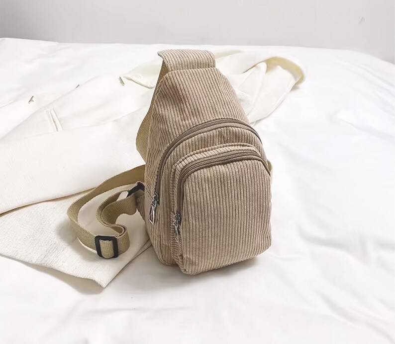 Puede incluir: Bolso bandolera de pana beige con un bolsillo frontal con cremallera y una correa ajustable. El bolso tiene una textura acanalada vertical y un peque&ntilde;o bolsillo con cremallera. La correa es de color beige claro con una hebilla negra.