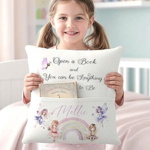 May include: White pillow with a pocket, featuring the text "Open a Book and You can be Anything to Be." Below the text is a pocket with the name "Mollie" and a rainbow design with fairies. A book is in the pocket.