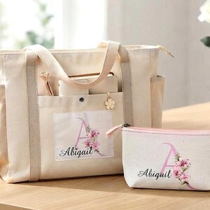 May include: A cream-colored canvas tote bag and a matching zippered pouch, both featuring a large pink letter A with floral accents and the name Abigail printed in black script. The tote has two sturdy handles and side pockets for storage.