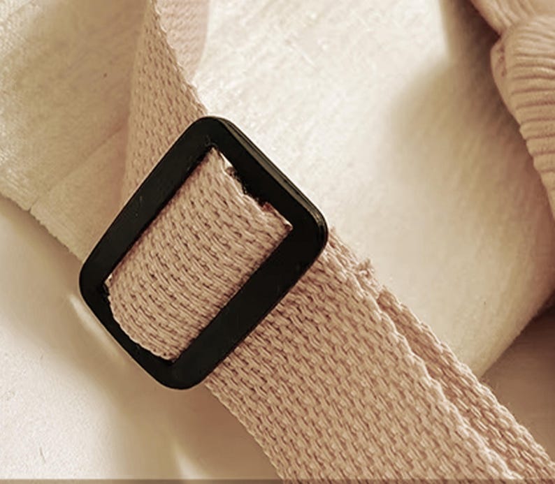 Puede incluir: Primer plano de una correa beige tejida con una hebilla cuadrada negra. La correa parece ser parte de un bolso o accesorio, con una superficie texturizada y una paleta de colores neutros.