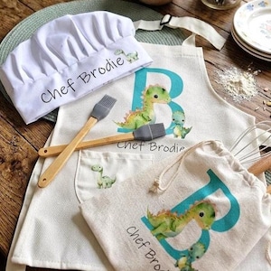 May include: A white chef's hat and apron set with a dinosaur theme. The apron features a large teal "B" and dinosaur illustrations. The set includes a matching drawstring bag and two wooden handled basting brushes. The text "Chef Brodie" is printed on the hat and apron.