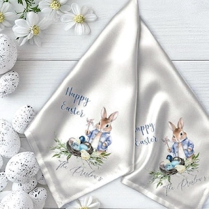 May include: Two white satin napkins featuring a watercolour illustration of a rabbit in a blue jacket holding a pinwheel next to a nest with blue eggs and a chick. The text reads Happy Easter and The Preslow's in a dark blue script font.