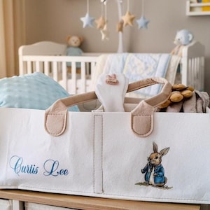 Puede incluir: Bolso tote de tela blanca con asas beige, bordado con el nombre "Curtis Lee" en escritura azul. Presenta una ilustración de Peter Rabbit. La bolsa está llena de artículos para bebés.