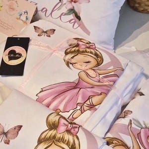 May include: A white fabric set featuring a repeating illustration of a ballerina with a blonde bun and pink bow, wearing a pink tutu. The design includes pink butterflies and soft floral accents. A black tag with a heart logo is attached to the fabric.