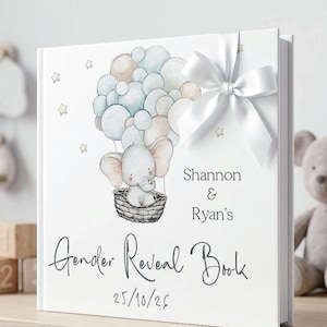 May include: A white hardcover book featuring a watercolor illustration of a baby elephant in a basket attached to a cluster of blue and beige balloons. The cover includes the text "Shannon & Ryan's Gender Reveal Book" and "25/10/26" with a white satin ribbon bow attached to the top right corner.