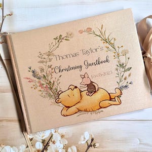 Puede incluir: Un libro de visitas beige con un diseño de corona floral y el texto "Thomas Taylor's Christening Guestbook 10-05-2023". La portada presenta una ilustración de oso y cerdo de dibujos animados. Una cinta beige está unida al libro.