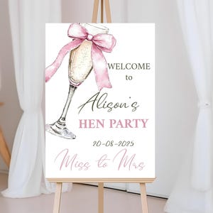 Może przedstawiać: Biały znak na sztaludze z ilustracją kieliszka do szampana i różową wstążką. Tekst brzmi "WELCOME to Alison's HEN PARTY 20-08-2025 Miss to Mrs."