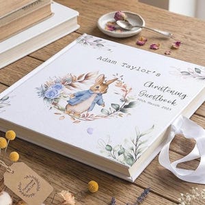 Puede incluir: Libro de visitas de bautizo blanco con una ilustración de acuarela de Peter Rabbit con una chaqueta azul, rodeado por una corona floral. El libro tiene el nombre "Adam Taylor" y la fecha "30 de marzo de 2023" impresos en la portada. Una cinta blanca está adjunta.