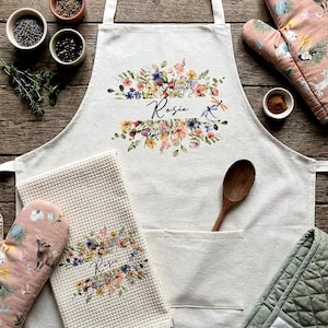 May include: Cream-coloured kitchen apron, oven mitt, and towel set with floral designs and the name "Rosie". The apron has a large pocket and a wooden spoon. The oven mitt and towel have a waffle texture.
