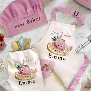 May include: A pink chef hat with the text Star Baker, a white apron with pink straps featuring a strawberry graphic and the text Star Baker Emma, and a matching white drawstring bag with the same strawberry design and the name Emma.