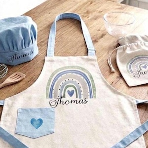 May include: A light-colored apron with a blue rainbow design and the name "Thomas" printed on it. A matching chef's hat and drawstring bag are also visible. The apron has a blue pocket with a blue heart.