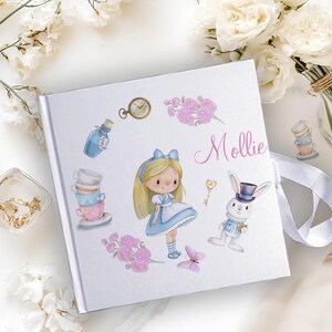 K&ouml;nnte beinhalten: Wei&szlig;es Fotoalbum mit einem blauen und wei&szlig;en Alice-im-Wunderland-Design. Das Album zeigt ein M&auml;dchen in einem blauen Kleid, ein wei&szlig;es Kaninchen, Teetassen und einen Schl&uuml;ssel. Das Album ist mit dem Namen "Mollie" personalisiert.