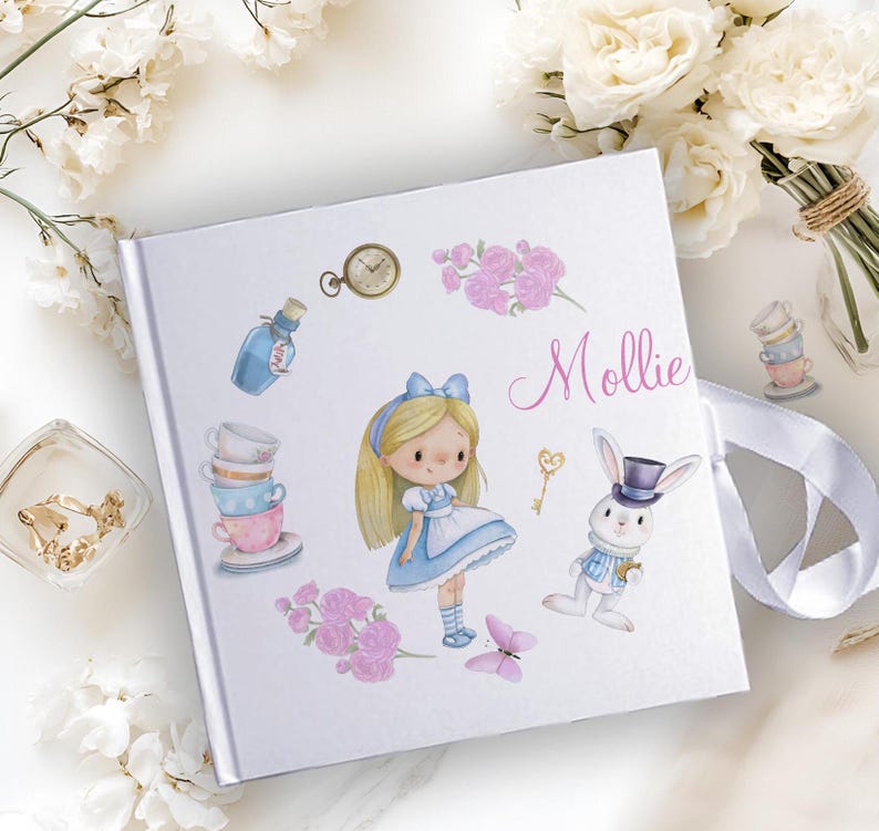 K&ouml;nnte beinhalten: Wei&szlig;es Fotoalbum mit einem blauen und wei&szlig;en Alice-im-Wunderland-Design. Das Album zeigt ein M&auml;dchen in einem blauen Kleid, ein wei&szlig;es Kaninchen, Teetassen und einen Schl&uuml;ssel. Das Album ist mit dem Namen "Mollie" personalisiert.