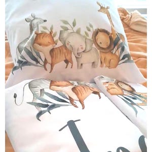 May include: A white pillowcase and matching bedding set featuring a watercolor illustration of a giraffe, lion, elephant, tiger, and deer surrounded by green leaves. The design includes a crescent moon and the name India printed in black script.