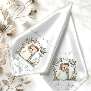 Puede incluir: Dos servilletas de satén blanco con una ilustración en acuarela de una pareja de novios bajo un arco de hojas. El texto dice Amelia & Thomas, Wedding Day y 03.08.2024. La tela tiene un acabado suave y reflectante con bordes limpios.