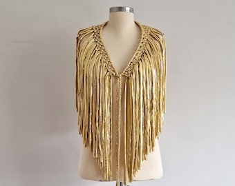 May include: A metallic gold fringe vest featuring a deep V-neckline and intricate macrame-style knotting along the collar. The garment is composed of long, flowing strips of shiny gold material that drape from the shoulders to the waist.