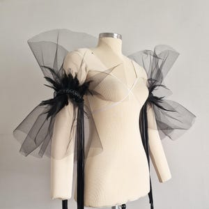 Może przedstawiać: Czarne tiulowe naramienniki z piórkowymi akcentami i długimi czarnymi wstążkami, prezentowane na manekinie. Tiul jest warstwowy, tworząc dramatyczny efekt. Pióra skupione wokół górnej części ramienia, dodając tekstury. Modny dodatek.