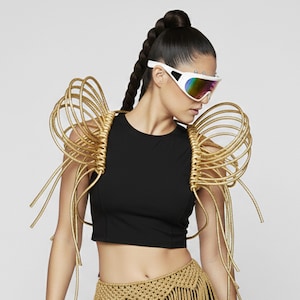 May include: A black sleeveless crop top featuring elaborate gold-colored coiled shoulder structures made of thick cord. The garment is paired with white-framed sunglasses with iridescent lenses and a gold-colored crocheted skirt with fringe.