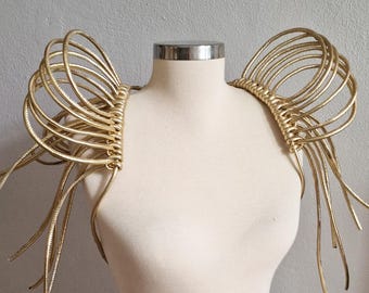 May include: A gold-colored shoulder accessory featuring multiple concentric circular loops that extend outward from the shoulders, with long, thin, flexible strands hanging down from the base of each circular structure on both sides.