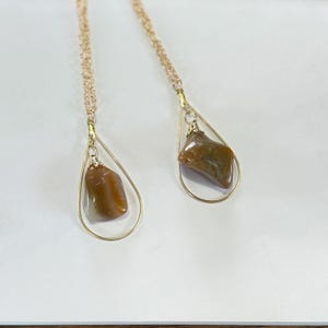 May include: Two gold-colored necklaces with teardrop-shaped pendants. Each pendant features a raw, brown gemstone suspended within a delicate gold wire frame. The necklaces rest on a white surface, showcasing the jewelry's design.