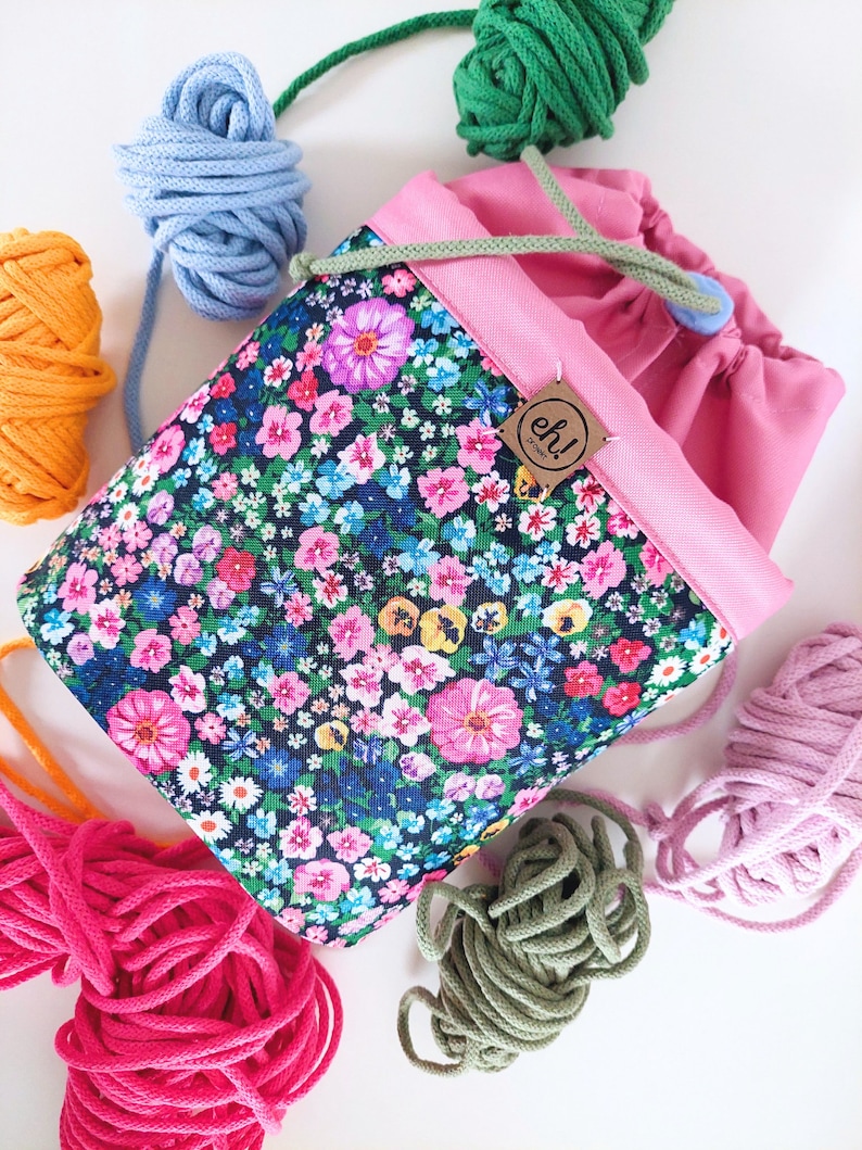 Pode incluir: Uma bolsa de projeto com cord&atilde;o e estampa floral com interior e acabamento rosa. A bolsa &eacute; cercada por meadas de fios coloridos em azul, laranja, rosa, verde e roxo. Uma pequena etiqueta com o texto "eh! project" est&aacute; anexada.