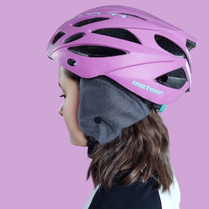 May include: A pink bicycle helmet with the word "meteor" in teal. The helmet has multiple vents and is worn with a gray ear warmer. The background is a solid light purple.