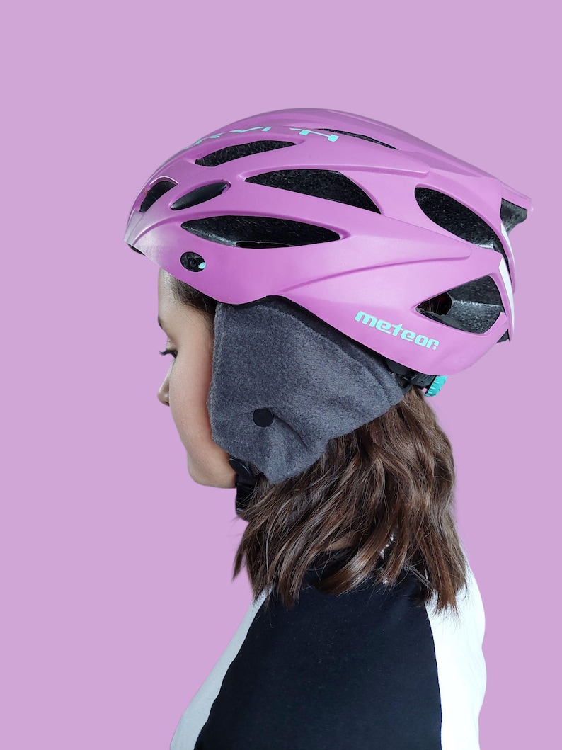 K&ouml;nnte beinhalten: Ein pinkfarbener Fahrradhelm mit dem Wort "meteor" in T&uuml;rkis. Der Helm hat mehrere Bel&uuml;ftungs&ouml;ffnungen und wird mit einem grauen Ohrenw&auml;rmer getragen. Der Hintergrund ist einfarbig hellviolett.