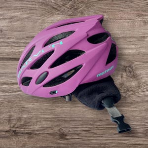K&ouml;nnte beinhalten: Ein magenta Fahrradhelm mit dem Wort "Meteor" auf der Seite. Der Helm hat mehrere Bel&uuml;ftungs&ouml;ffnungen und einen schwarzen Kinnriemen mit Schnalle. Auf der Oberseite des Helms steht das Wort "mivi" in hellblauer Farbe.