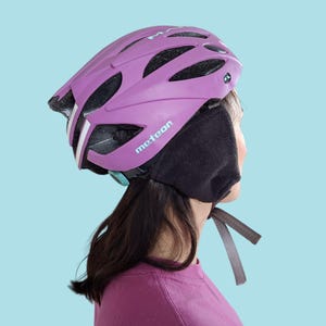 K&ouml;nnte beinhalten: Ein lila Fahrradhelm mit wei&szlig;en Streifen und dem Wort "meteor" in Wei&szlig;. Der Helm wird mit schwarzen Ohrenw&auml;rmern getragen. Der Hintergrund ist hellblau.