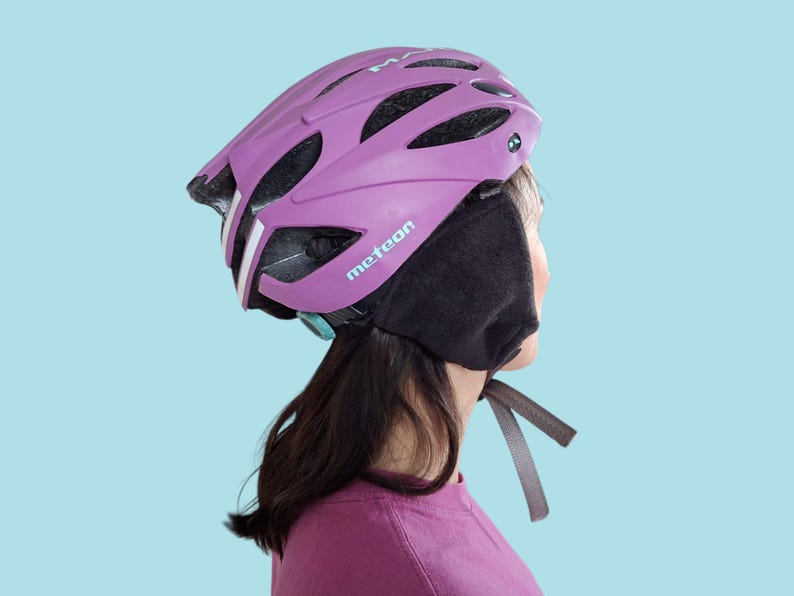 K&ouml;nnte beinhalten: Ein lila Fahrradhelm mit wei&szlig;en Streifen und dem Wort "meteor" in Wei&szlig;. Der Helm wird mit schwarzen Ohrenw&auml;rmern getragen. Der Hintergrund ist hellblau.