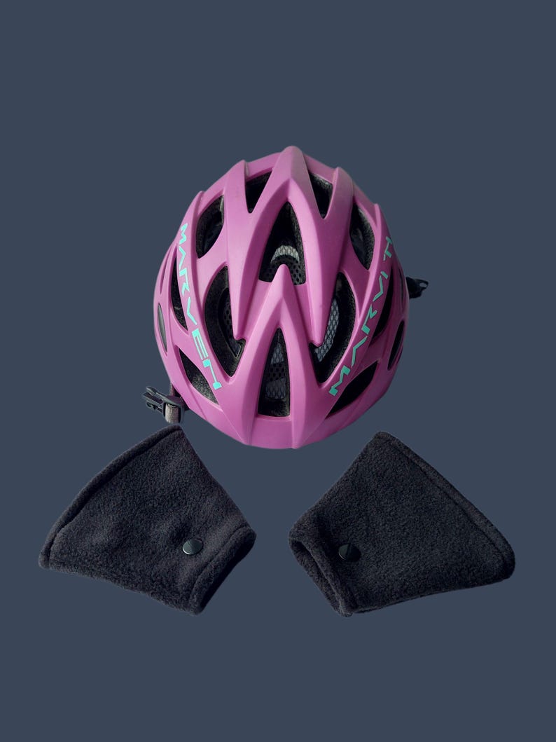 K&ouml;nnte beinhalten: Ein magenta Fahrradhelm mit dem Wort "MARVET" in t&uuml;rkisfarbenen Buchstaben. Der Helm hat mehrere Bel&uuml;ftungs&ouml;ffnungen und ein schwarzes Innenfutter. Zwei schwarze Ohrensch&uuml;tzer mit Druckkn&ouml;pfen sind enthalten.