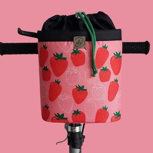 May include: A pink handlebar bag with a black drawstring top and a green drawstring. The bag is covered in a white and red strawberry pattern. The bag is attached to a black handlebar.