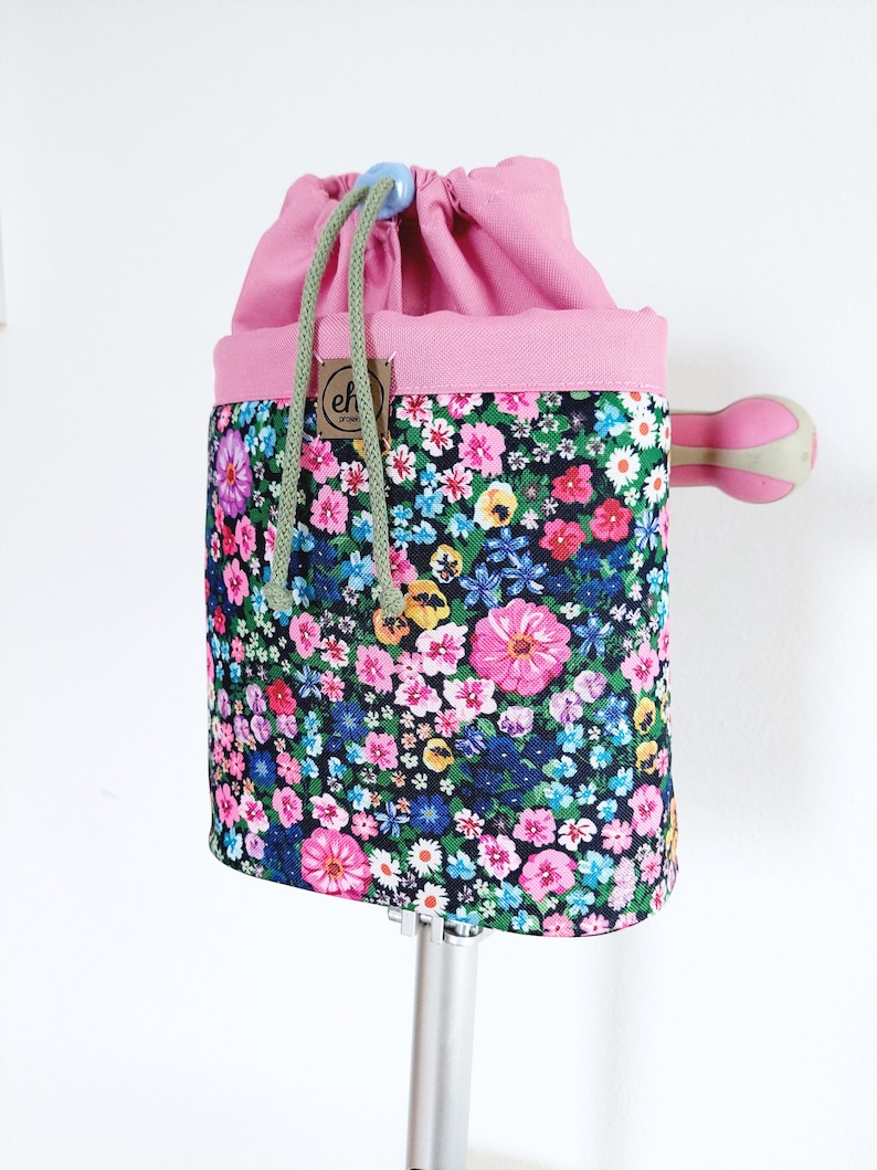 Pode incluir: Uma bolsa com cord&atilde;o floral com uma parte superior rosa e um cord&atilde;o verde. A bolsa apresenta um padr&atilde;o floral colorido com flores rosa, azuis, amarelas e brancas sobre um fundo escuro. Uma pequena etiqueta castanha com as letras "eh" est&aacute; anexada.