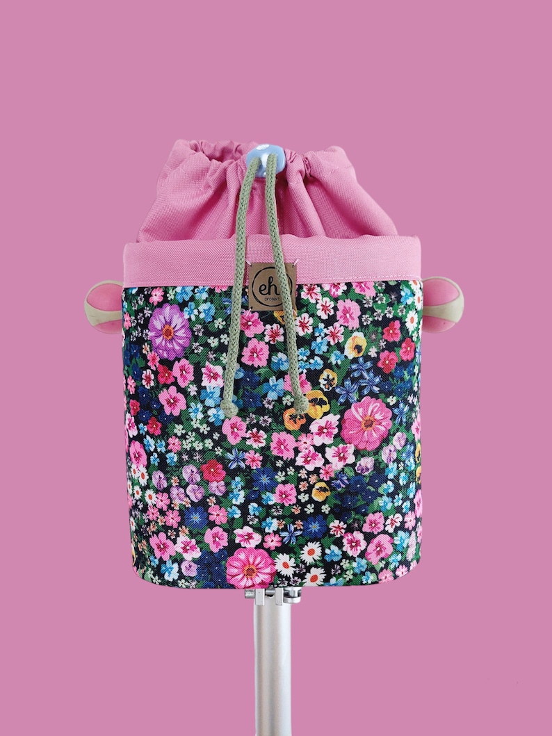 Pode incluir: Uma bolsa com cord&atilde;o floral com a parte superior e acabamento rosa. A bolsa apresenta um padr&atilde;o colorido de flores rosa, azuis e amarelas sobre um fundo preto. O cord&atilde;o &eacute; verde e a bolsa tem uma pequena etiqueta com as letras "eh".